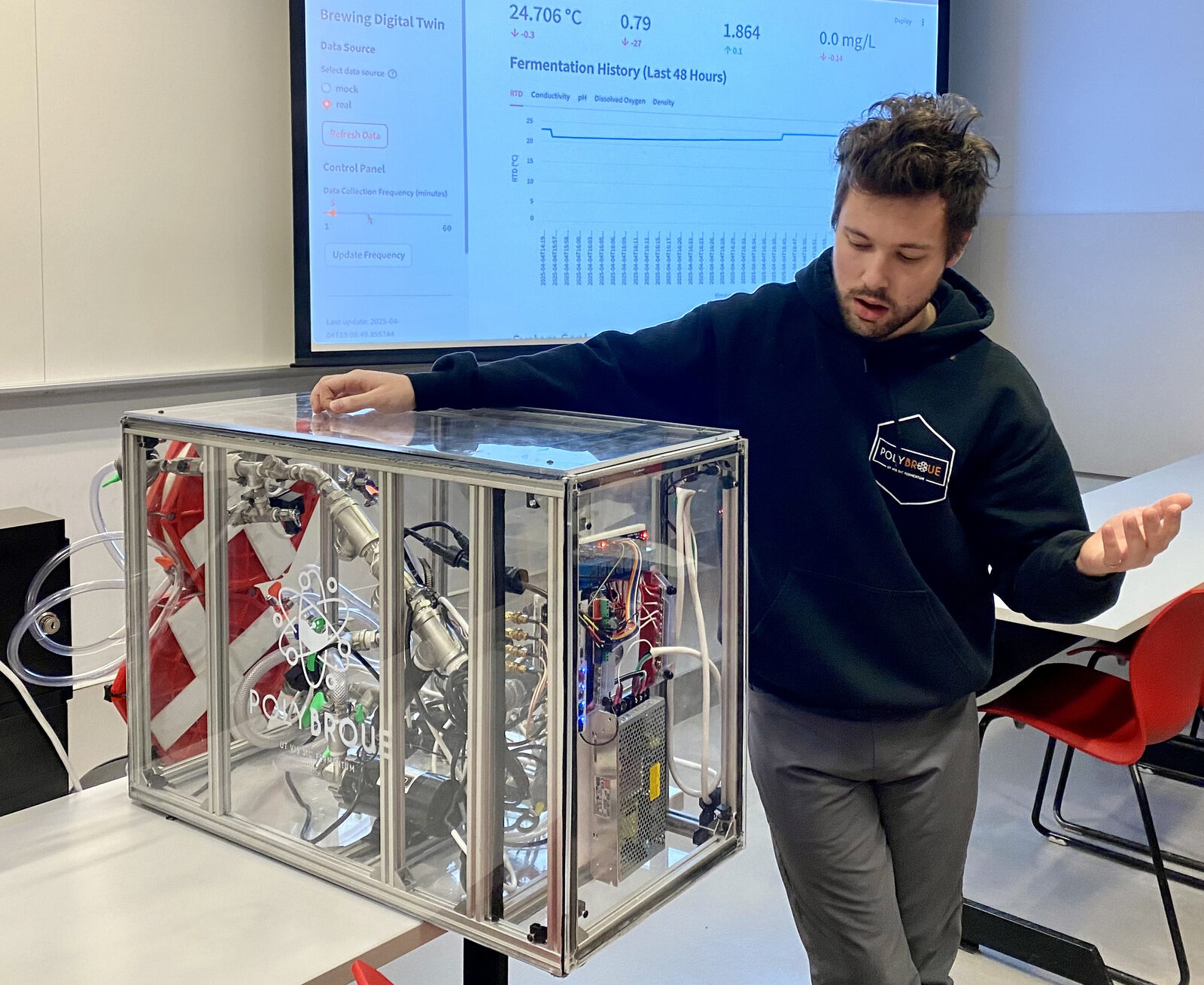Raphaël Tremblay, wearing a Polybroue hoodie, stands beside the sampling equipment in a clear acrylic case, with the digital twin dashboard showing fermentation history on the screen behind him.